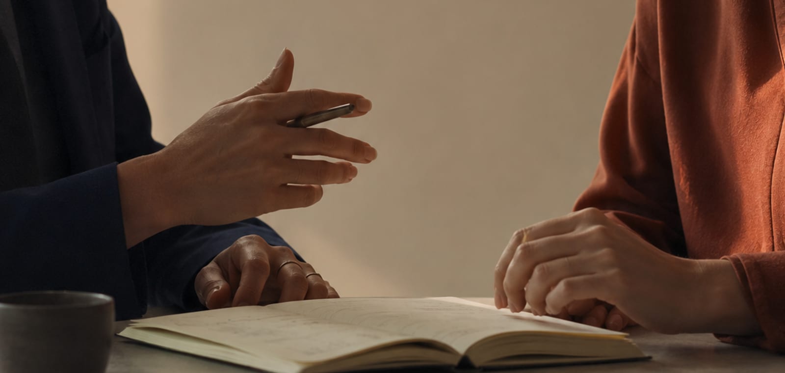 Duas pessoas sentadas à mesa, mãos sobre um caderno aberto durante uma conversa de trabalho