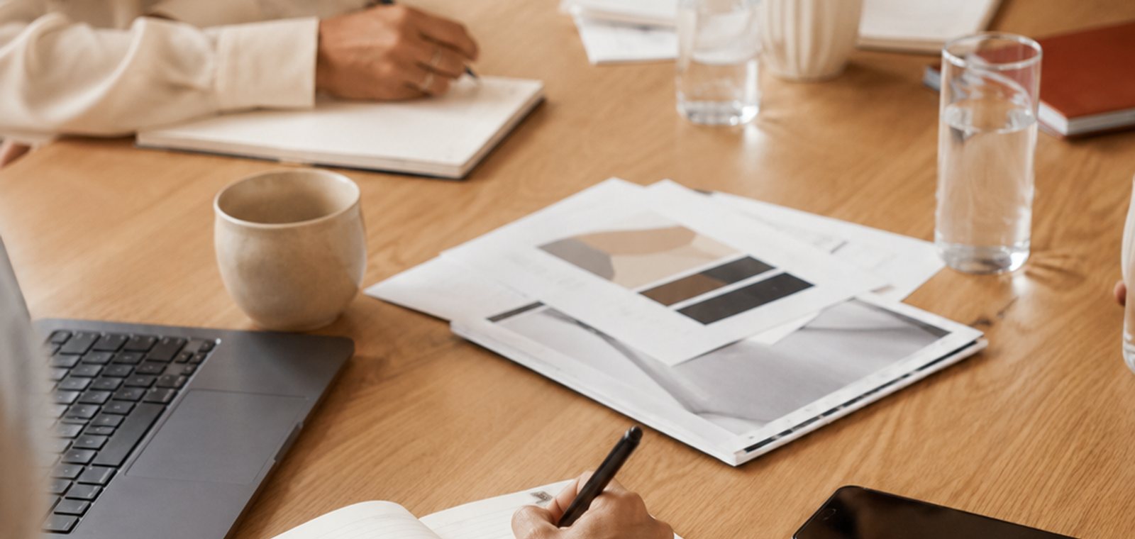 Mesa de reunião com caderno, papéis e laptop durante uma conversa de trabalho